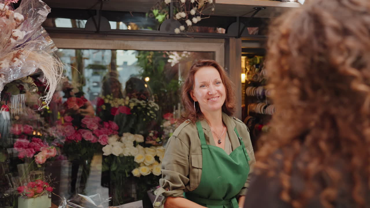 Fiorista accoglie una cliente con i capelli ricci al bancone con un bouquet e sorrisi, fiorista offre consulenza e consigli, grembiule verde, sfondo di boutique affollata piena di rose e nastri regalo