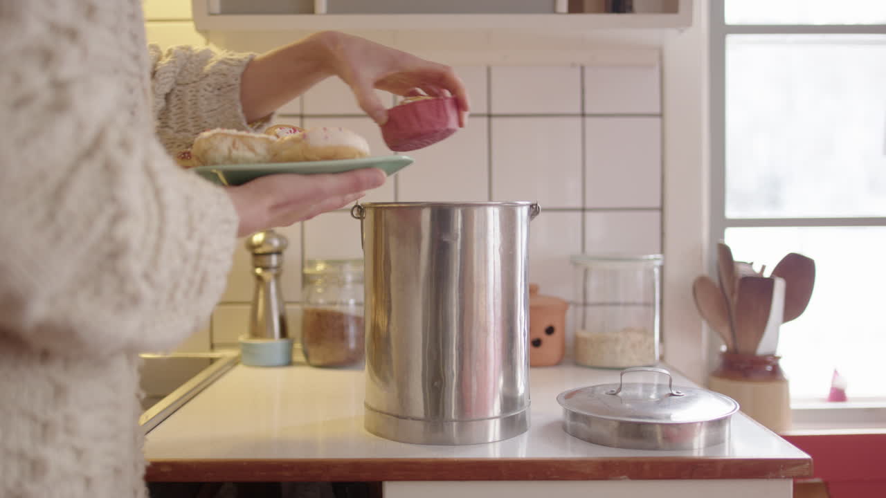 una mujer lanza rosquillas viejas y rancias a un contenedor de compost de acero en la cocina.