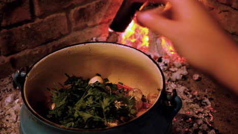 adding herbs to dish being cooked in cast iron pot on open fire