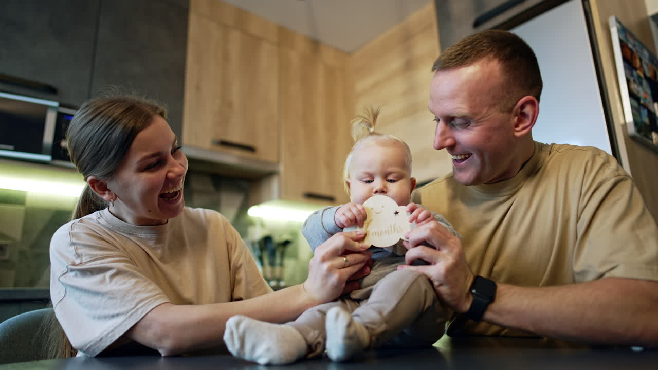 Baby of seven months sits on the table. Happy parents rejoice and kiss the cute child. Low angle view.