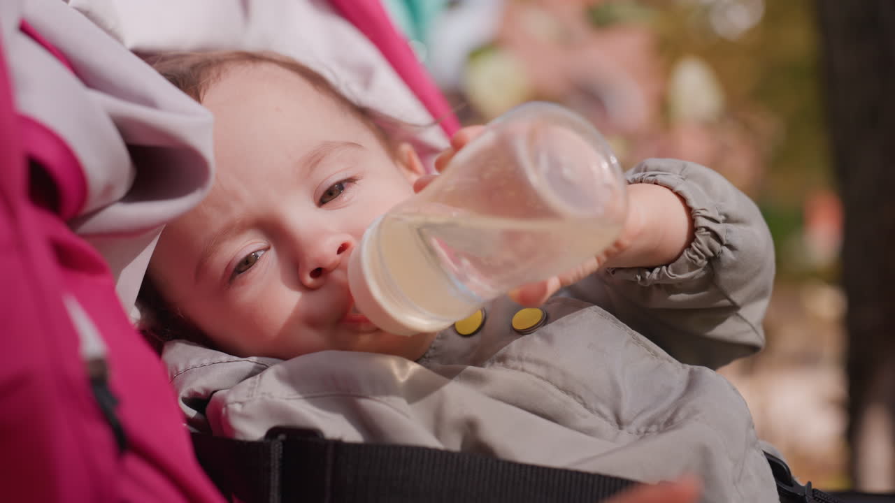Neonato nel passeggino che beve tranquillamente dal biberon all'aperto al parco, primo piano dell'espressione dolce e del lento sorso, tettuccio rosa e brezza leggera, tono rilassante di una gita in famiglia con luce diurna serena e cura amorevole.