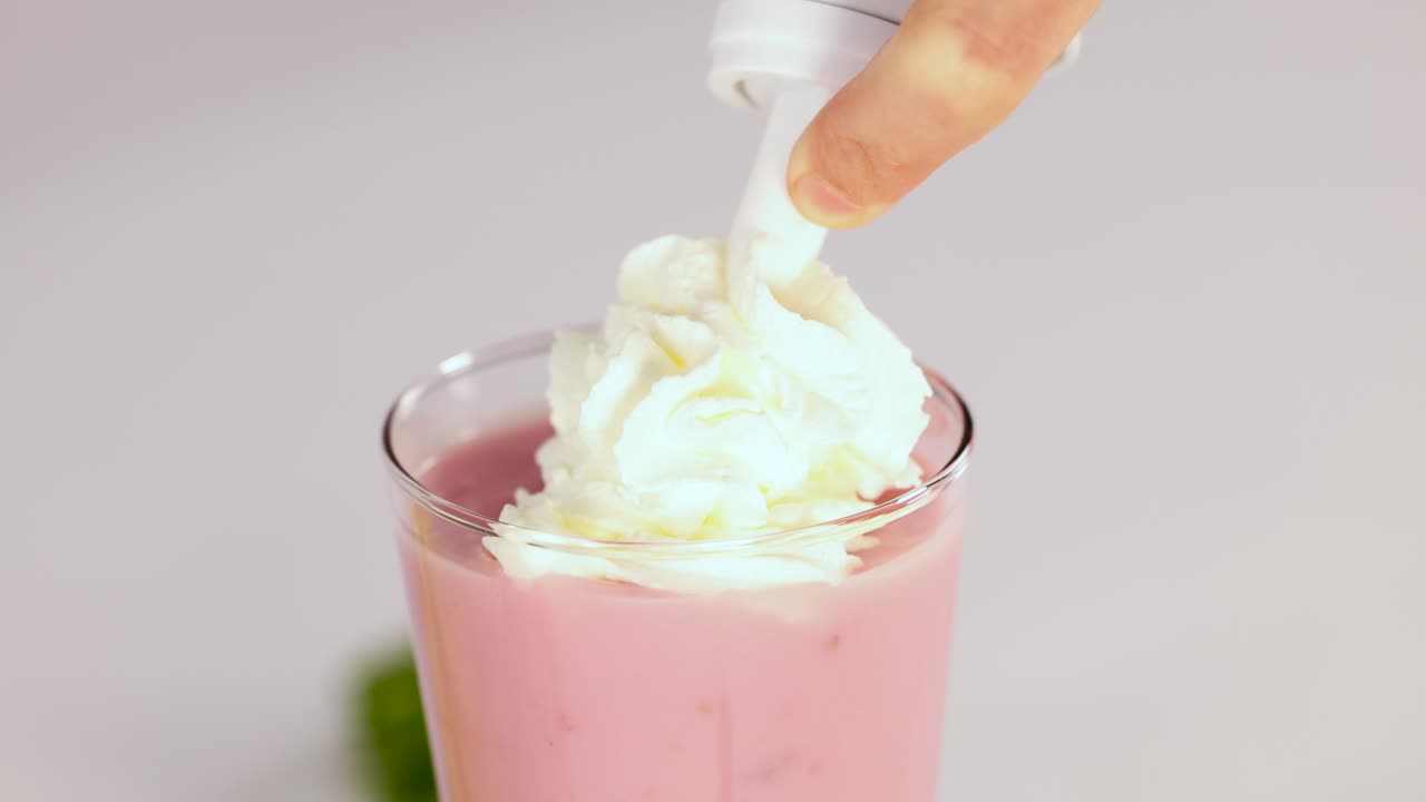Hand dispenses whipped cream onto pink strawberry milkshake in glass, bright lighting, close-up view