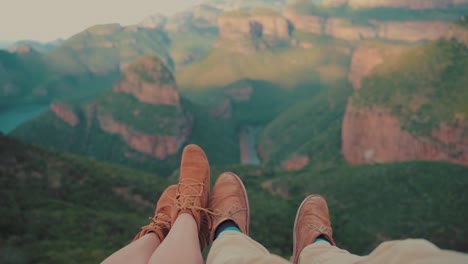 Video-shot-of-feet-of-a-couple-sitting-on-the-edge-of-a-cliff,-looking-over-the-valley-and-vast-canyon