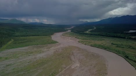 scenic panoramic summertime flight above yukon kluane brown river by evergreen trees and green vegetation towards mountain range, dramatic clouds, rain and grey sky, canada, overhead aerial approach