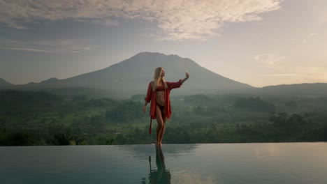 fit attractive blond woman in red bikini standing on infinity pool edge taking selfie, mount agung volcano in background, sunrise