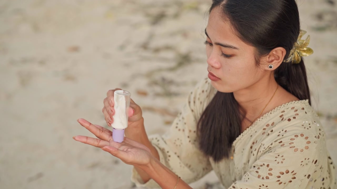 Asian woman on beach checking forehead then dispensing sunscreen spray into palm, inspecting skin for redness, pensive expression with hair tied by flower clip, closeup of bottle and mist, protective