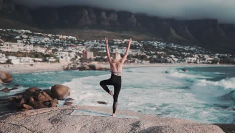 hermosa y saludable mujer haciendo una pose de yoga frente a un increíble paisaje marino azul con montañas nubladas en la parte de atrás