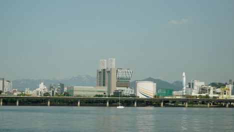 Pequeño-Yate-Con-Velas-Plegadas-Navegando-Por-El-Río-Hangang-Con-La-Autopista-Gangbyeon-Y-El-Distrito-De-Mapo-gu-Del-Centro-De-Seúl-Al-Fondo,-Las-Montañas-Bukhansan-Detrás-Del-Paisaje-Urbano