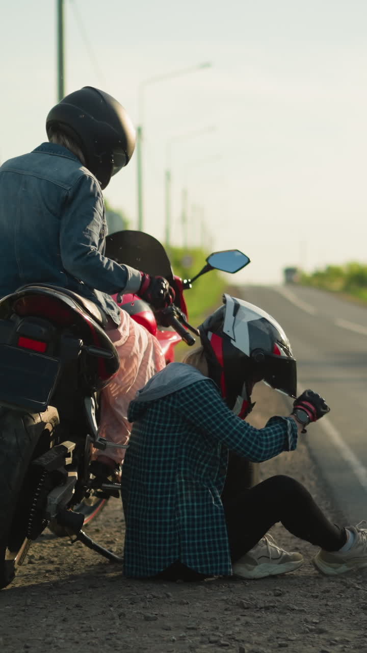 도로 에 있는 두 명의 여성, 한 명은 오토바이에 앉아 옷을 조정하고 다른 한 명은 바닥에 누워 헬 비저를 닫고, 둘 다 도로  에 있습니다.