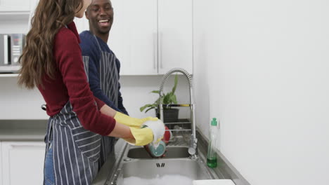 una pareja feliz y diversa lavando platos, hablando y riendo en la cocina, en cámara lenta.