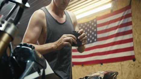 a male mechanic in a gray t-shirt takes out a metal part from the back of his motorcycle in a garage - workshop. workshop with a us flag and various tools for servicing equipment