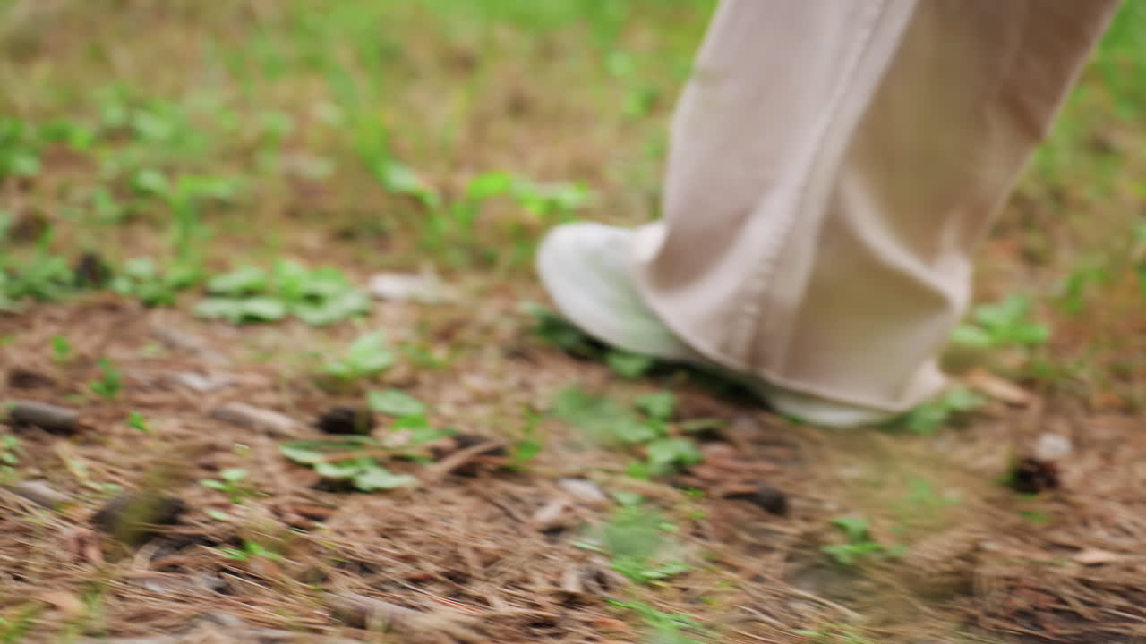 마른 잎와 푸른 풀로 덮인 숲길을 운동화를 신고 천천히 걷는 소녀의 다리를 클로즈업한 장면이에요. 평화로운 날 자연 속에서 자연스러운 야외 활동, 차분한 분위기, 자연과의 교감을 보여줘요.