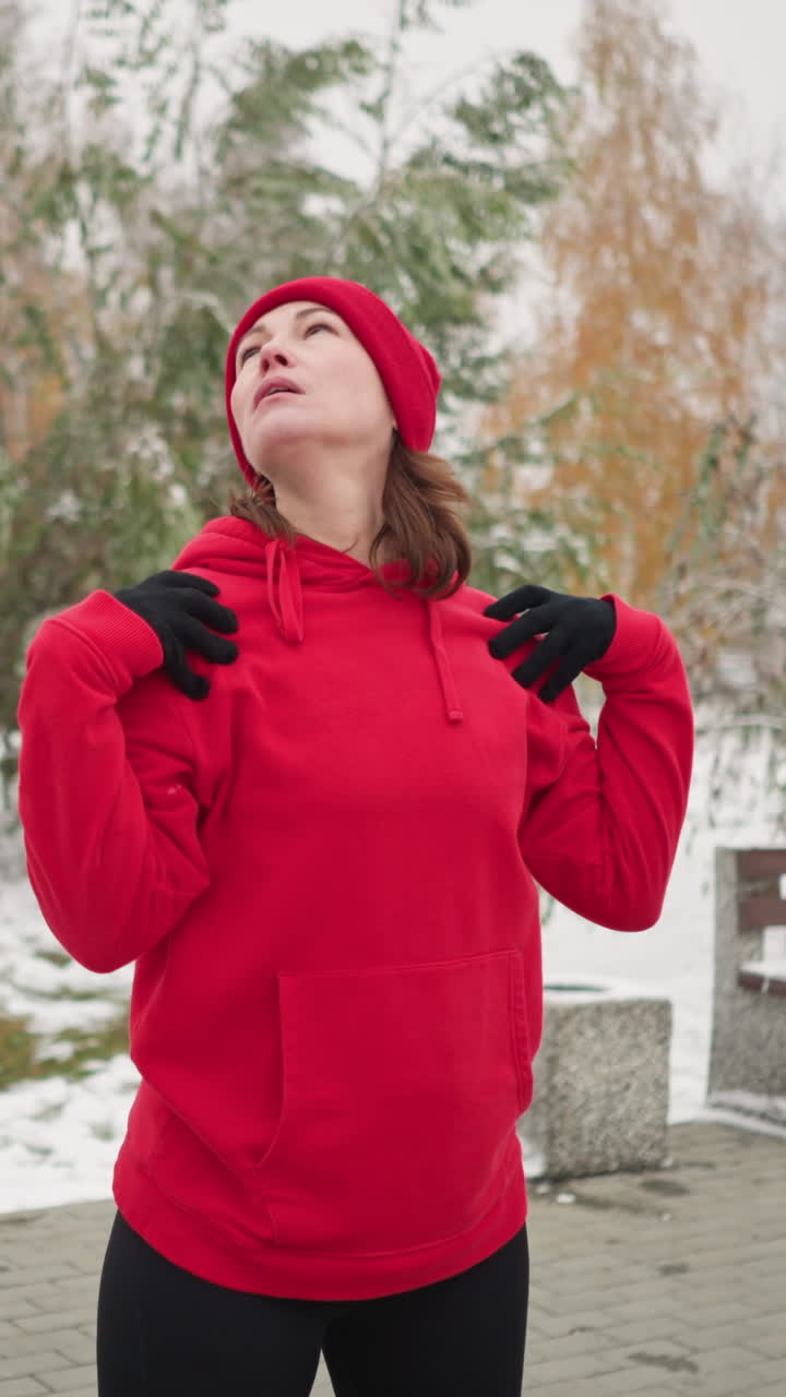 coach in red beanie stands outdoors with hands on shoulders, performing neck stretches by turning head left and right in serene winter setting with snow-covered ground, benches, and trees