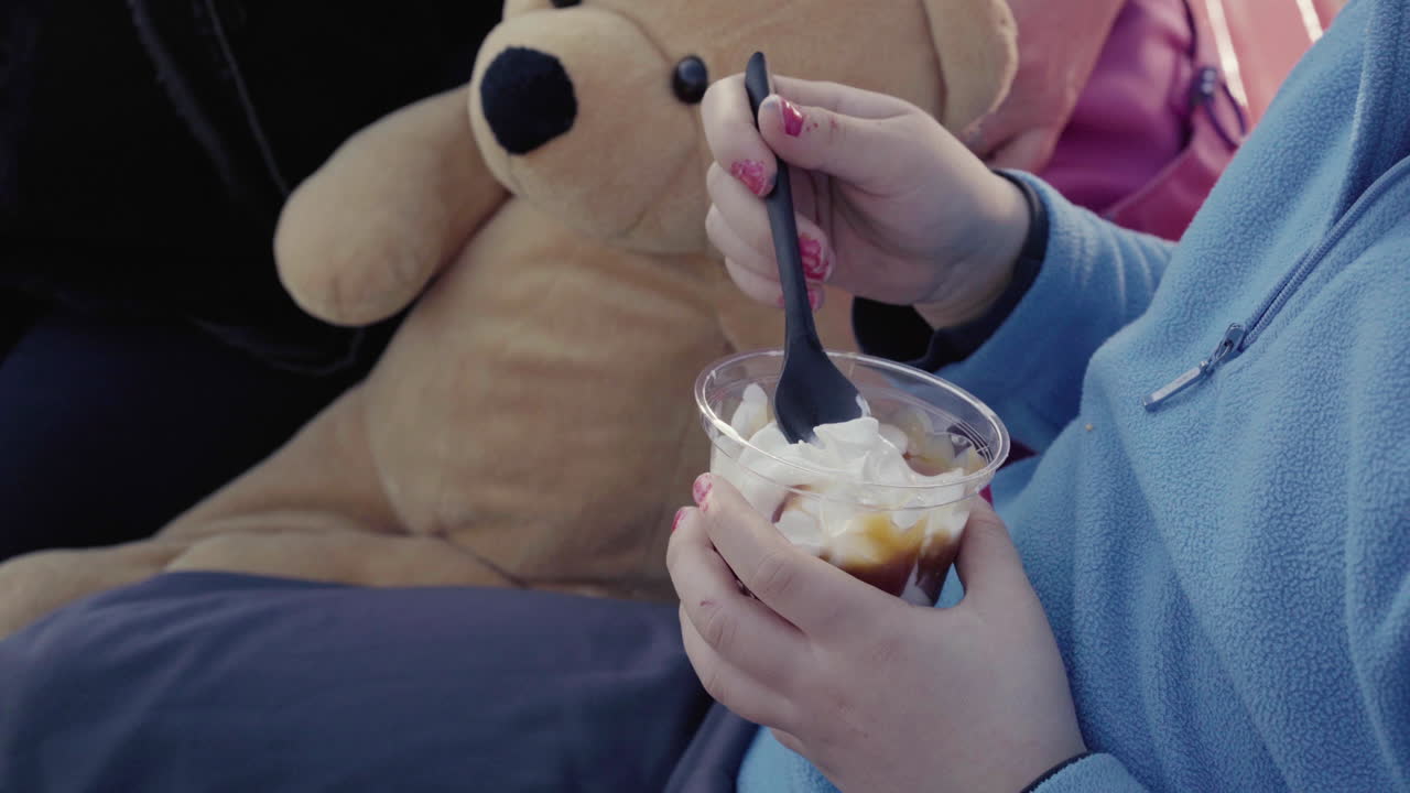 파란색 점퍼 매니큐어와 테디베어를 들고 야외에 앉아 있는 선디 아이스크림을 들고 있는 어린 소녀의 클로즈업