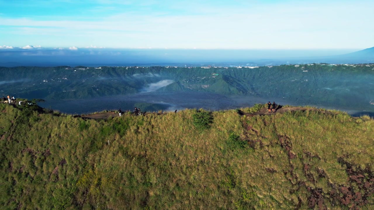 Punto panoramico montano che si affaccia sulla caldera del Monte Batur con escursionisti che esplorano un sentiero panoramico sulla cresta sopra un ampio bacino craterico vulcanico e gli aspri altopiani di Bali durante la morbida luce del mattino, ripresa laterale con drone