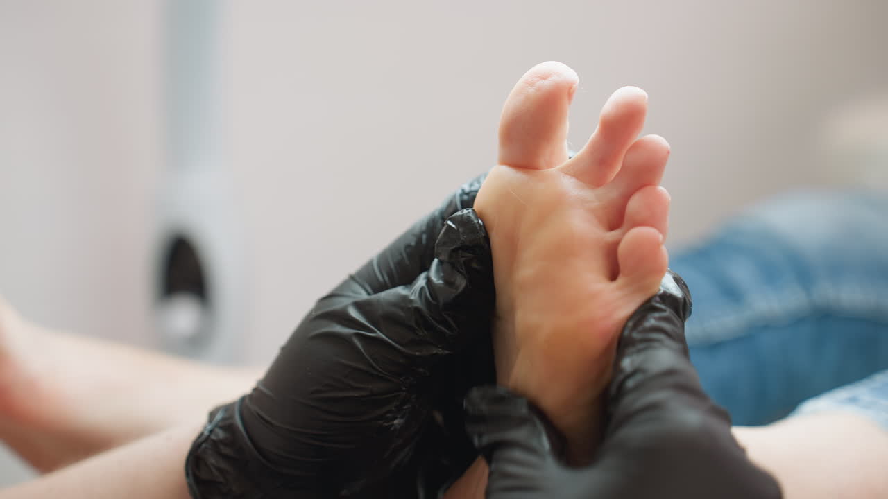 Close-up view of technician wearing black gloves gently massaging underside of client foot during wellness treatment. Client leg in denim visible as technician focuses on sole area under bright light