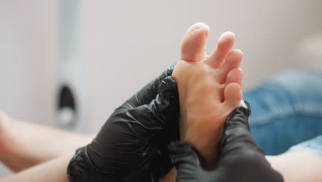 close-up view of technician wearing black gloves gently massaging underside of client foot during wellness treatment. client leg in denim visible as technician focuses on sole area under bright light