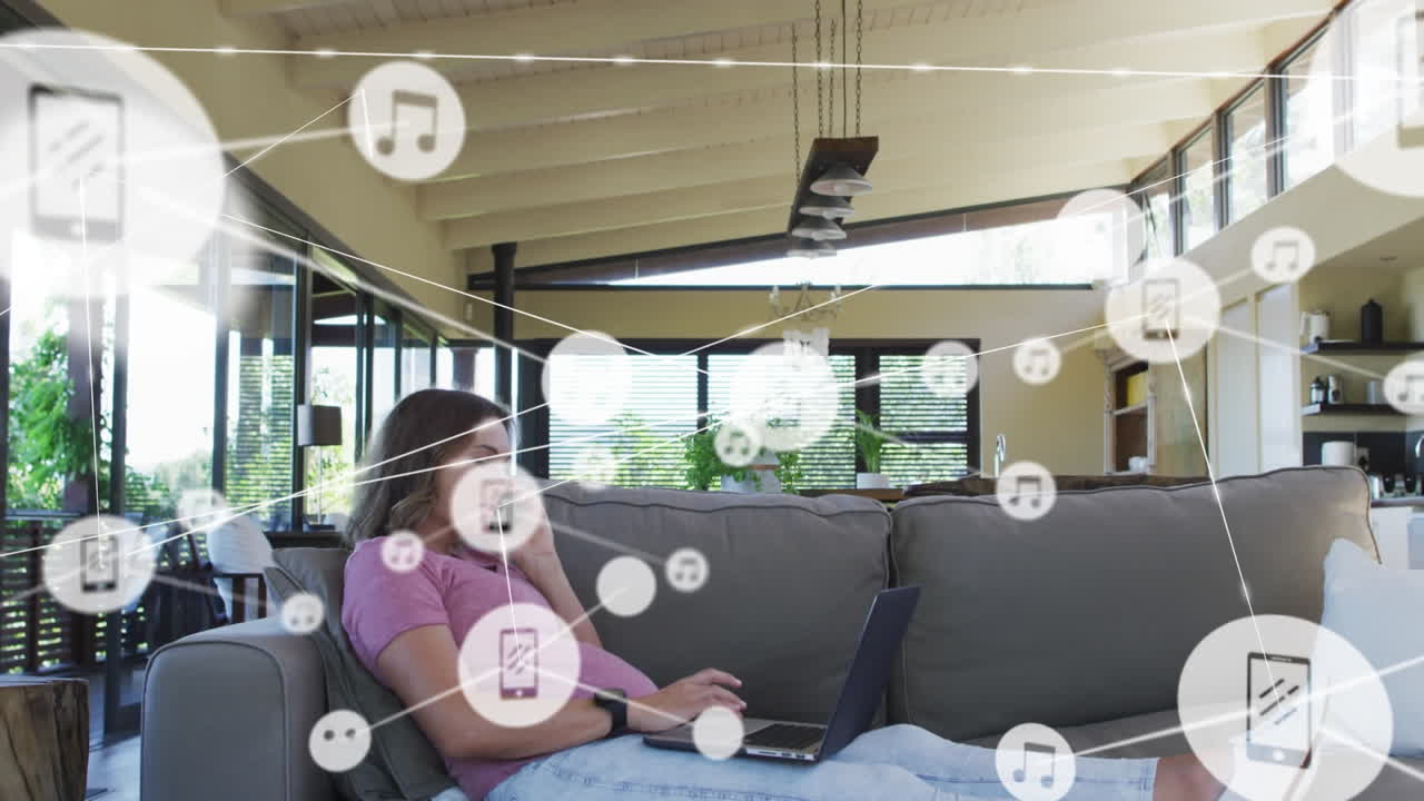 animación de iconos conectados, mujer caucásica sentada en el sofá hablando por teléfono mientras usa una computadora portátil