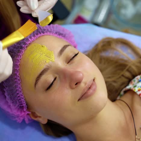 cosmetologist applying gold mask on woman's face using brush in cosmetology. portrait of young girl on cleaning skin procedure by beautician in beauty clinic. video from the top