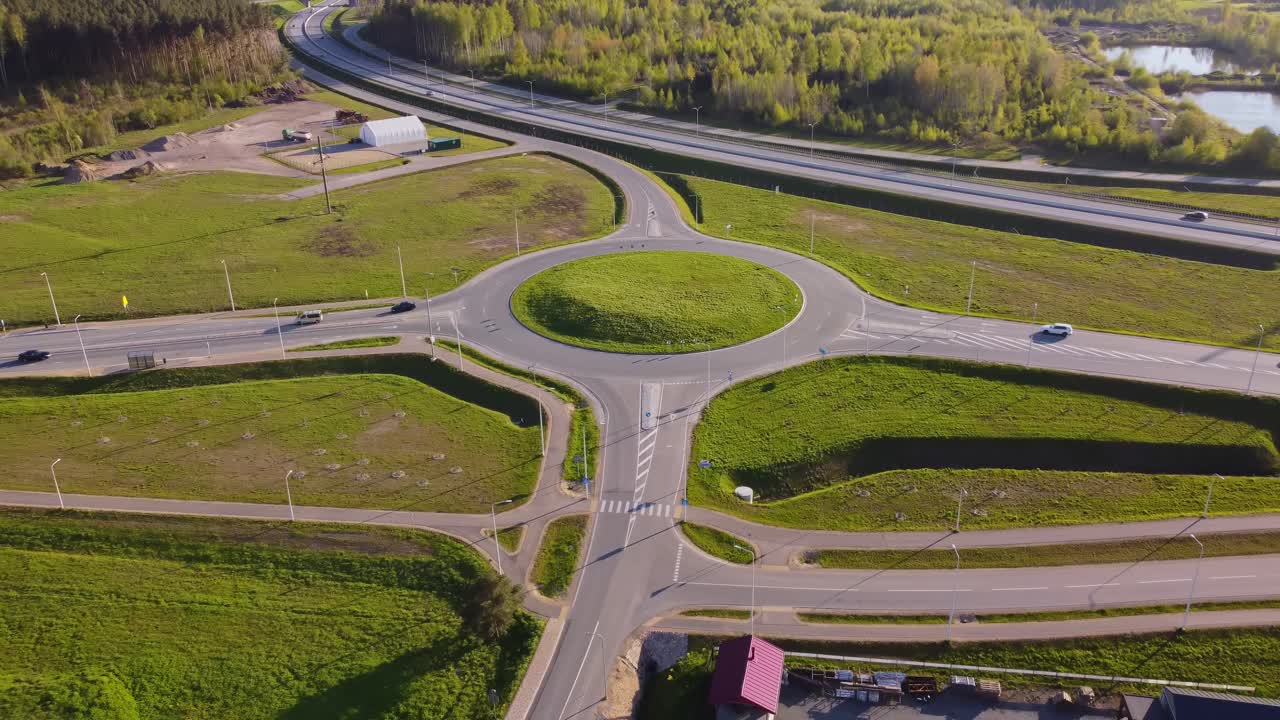 Drone pulls out revealing a roundabout at Katlakalns with grassy center, intersecting roads, adjacent fields, and distant highway bordered by forest