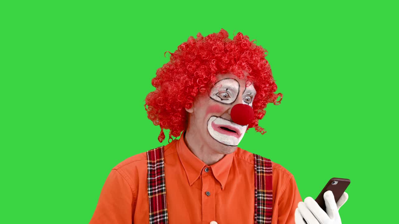 payaso gracioso mirando el teléfono inteligente y reaccionando a lo que ve en una pantalla verde, clave de croma