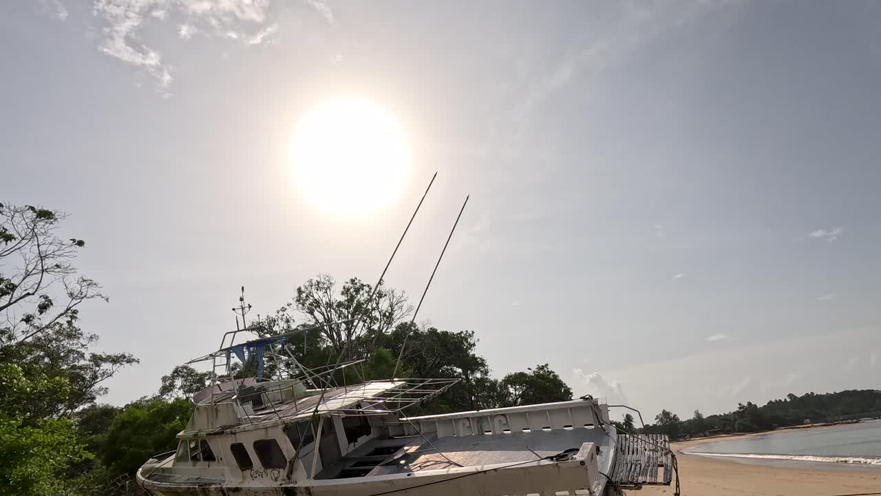 A stationary camera slowly pans to reveal an abandoned boat beached on sand under bright midday sun, with lush greenery and calm water nearby