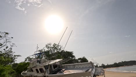 a stationary camera slowly pans to reveal an abandoned boat beached on sand under bright midday sun, with lush greenery and calm water nearby