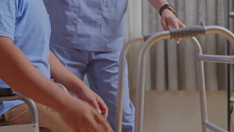 아시아 남성 간호사가 체어에 앉아있는 환자를 도와주고, 집에서 물리 치료를 하는 동안 스스로 서서 걸을 수 있는 워커를 사용하고 있다.