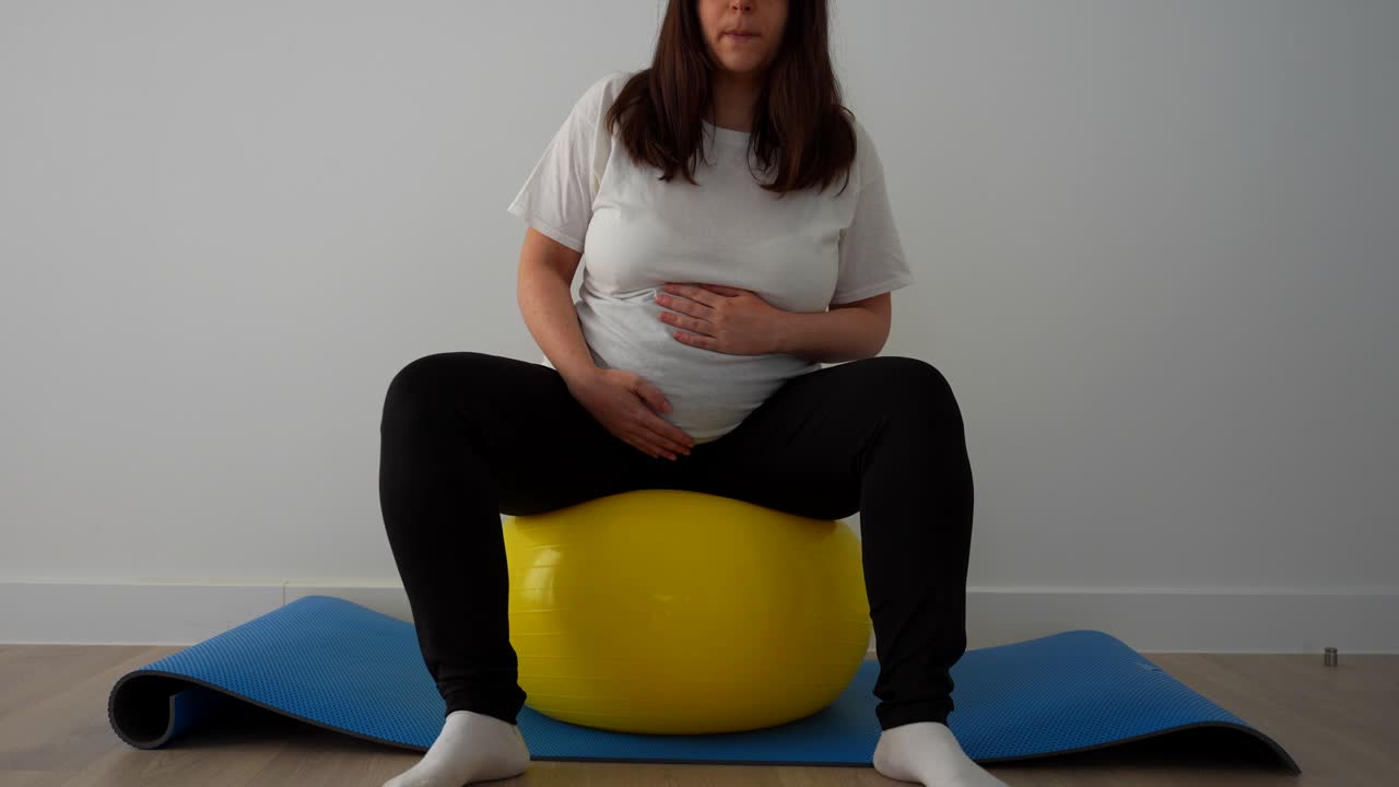 Pregnant woman doing delivery prep circles on a yellow fitball at home on a mat