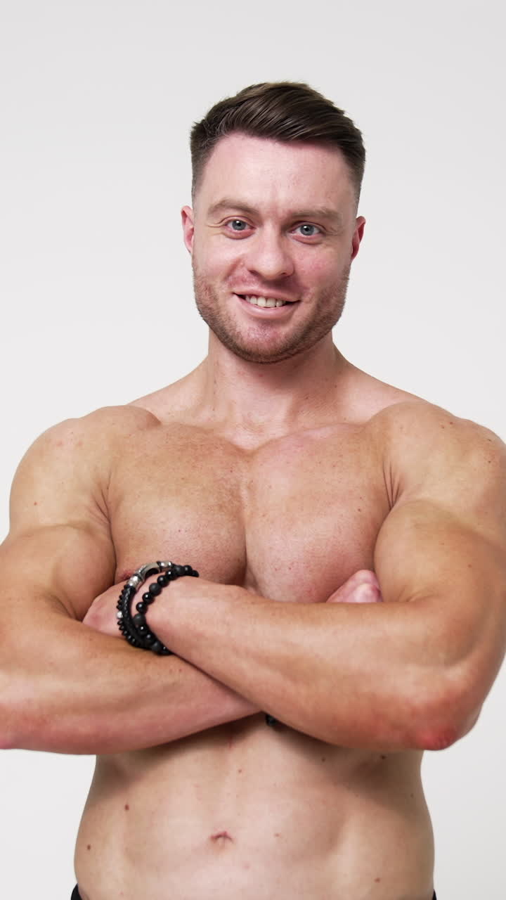 Sportive man with naked torso holding hands in pockets of black trousers. Man crosses hands on chest smiling to camera. White backdrop. Vertical video