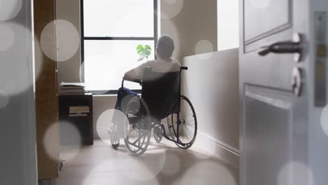 animation of light spots over senior biracial man sitting in wheelchair