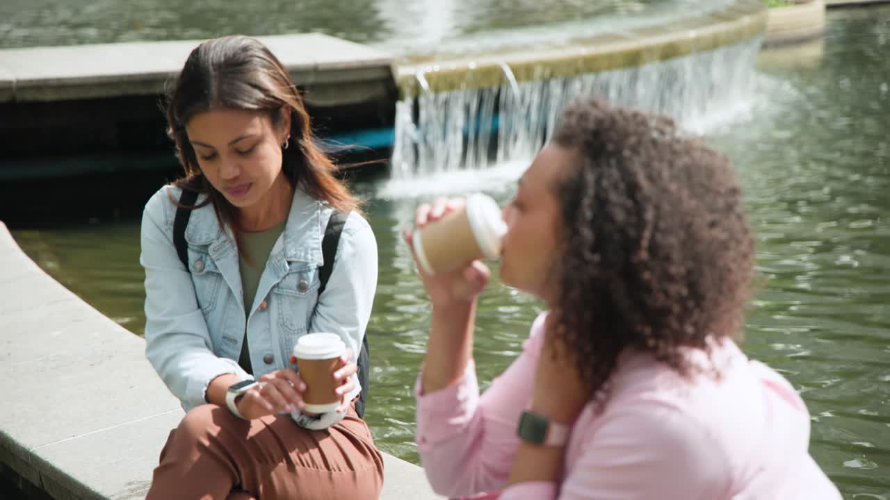 African American women sitting by pond right woman turning and speaking sipping to-go cups chatting