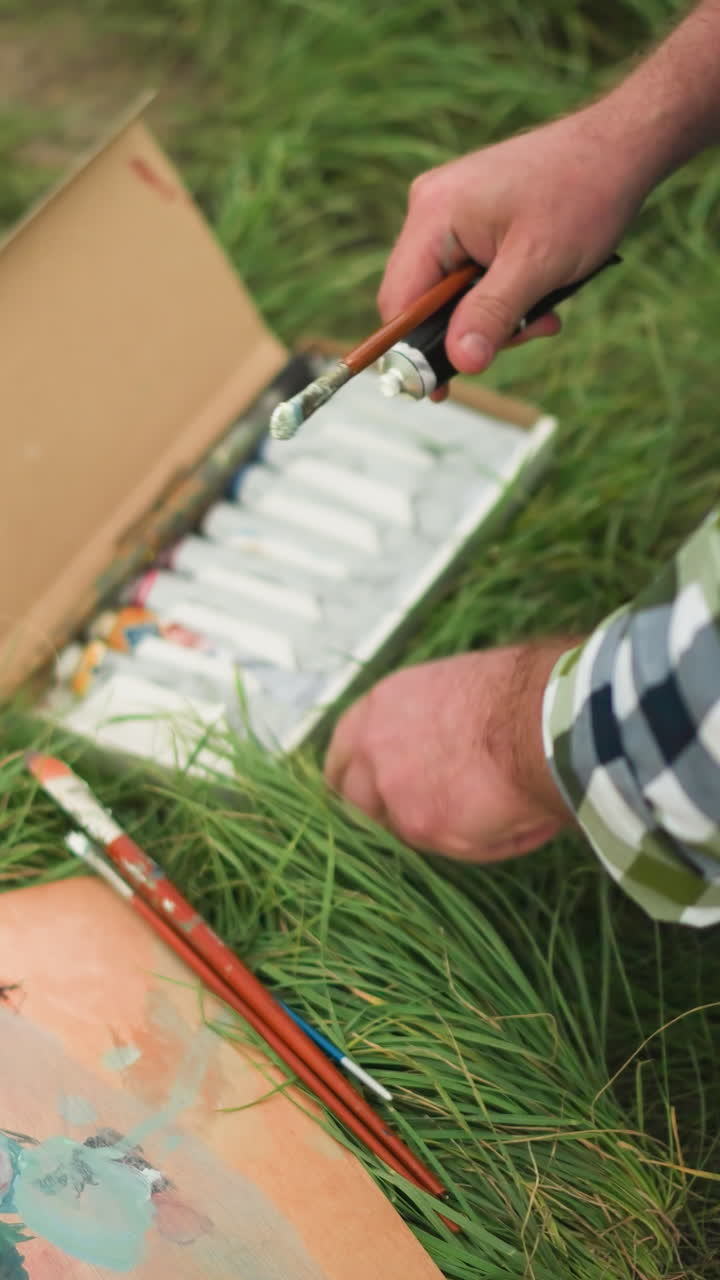 An artist in a checkered shirt bends down in a grass field, carefully opening a paint tube from a box of paints. The scene captures the preparation stage of outdoor painting
