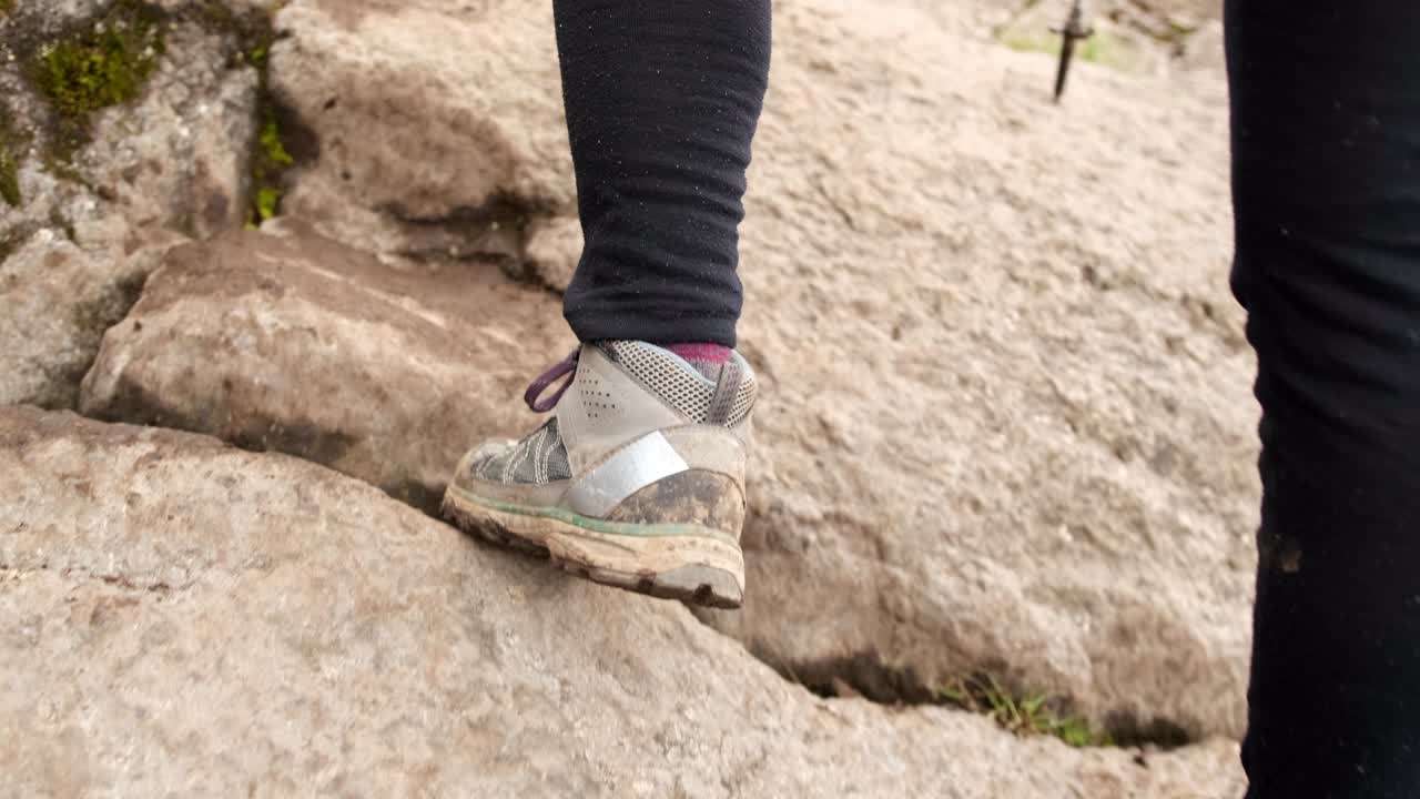 Rear view of a female tourist's leg in trekking boots climbing up the rocks with trekking poles.