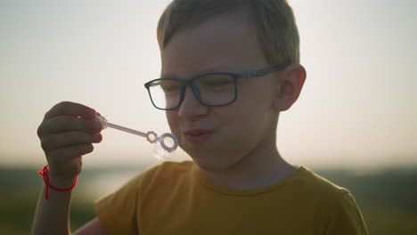 primer plano de un niño con una camisa amarilla y gafas mientras sumerge una varita en una botella de espuma y sopla burbujas en el aire al atardecer, capturando la alegría e inocencia de la infancia