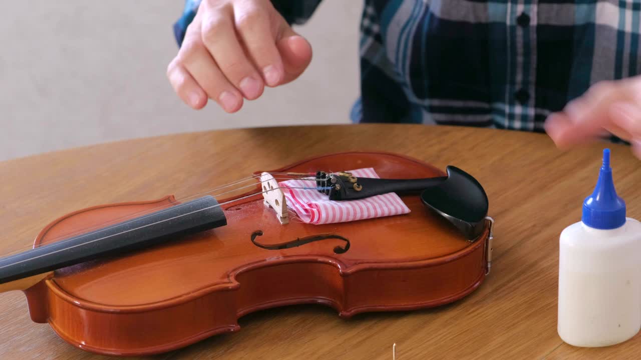 Close-up hands of young man in plaid shirt is repairing a violin sitting at the table.