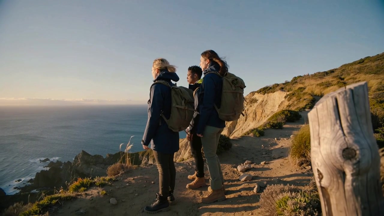 Approaching lookout, three hikers pausing at cliff edge, watching ocean with jackets and packs