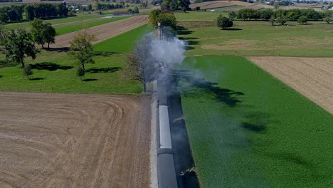 an aerial view from directly above a steam train blowing smoke and steam at the drone as it travels thru the countryside