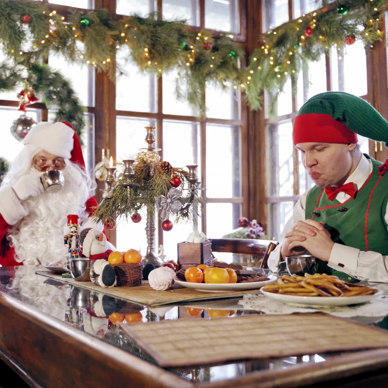 Two funny elves and Santa sitting at festive table on Christmas background. Girl elf tries to take sweets and another elf interferes her.