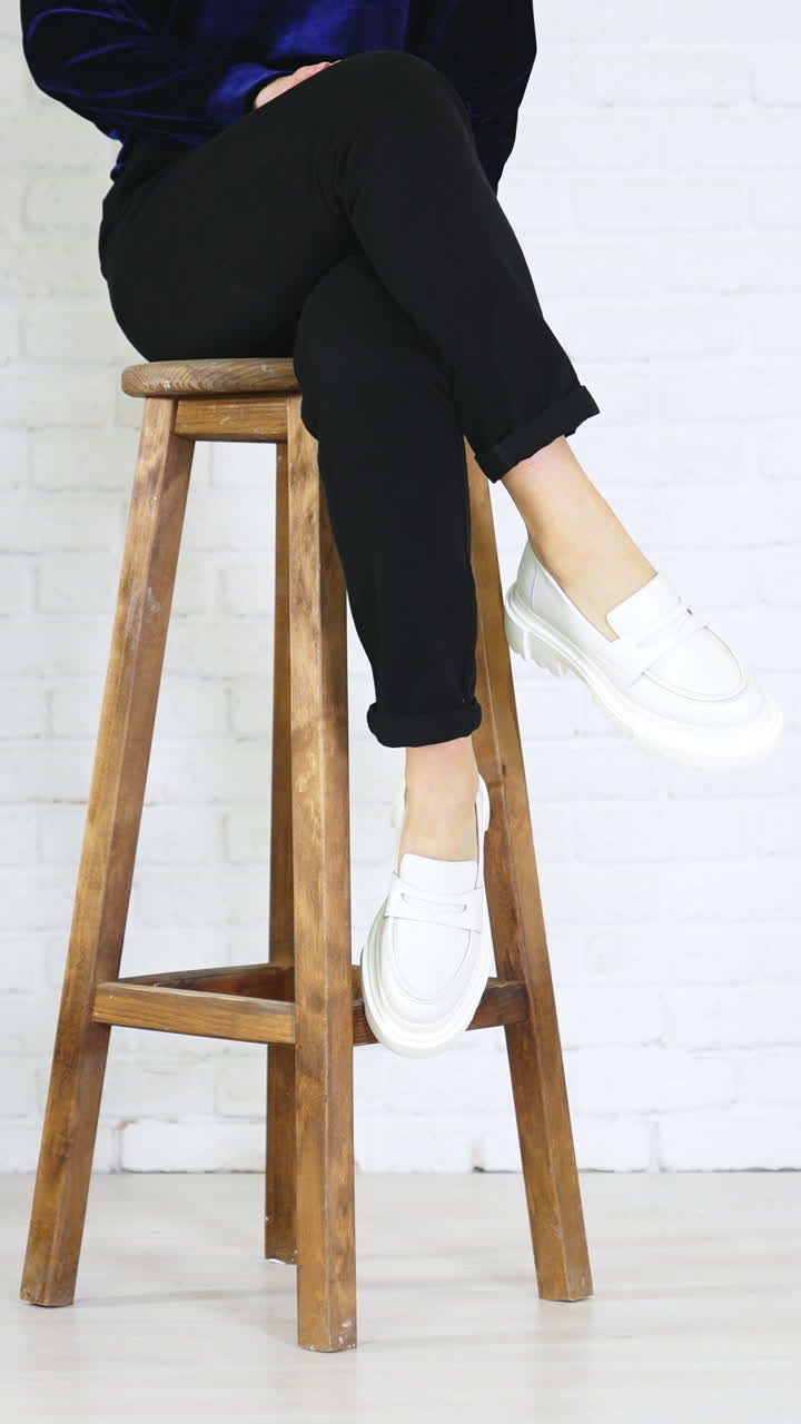 Unrecognized woman sitting on the stool showing her new shoes. Presenting white modern design footwear at white wall backdrop. Vertical video