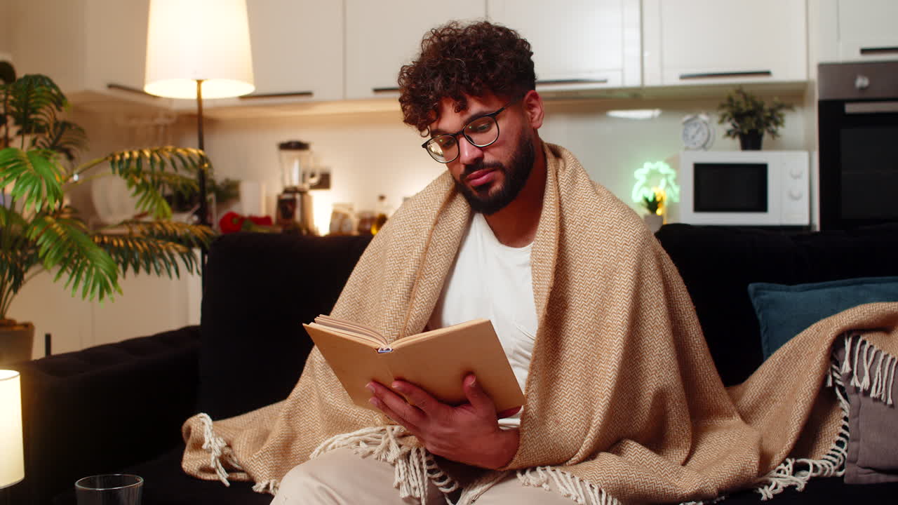 Arabian man in glasses reads favorite book wrapped in blanket quiet evening relax calm focus night