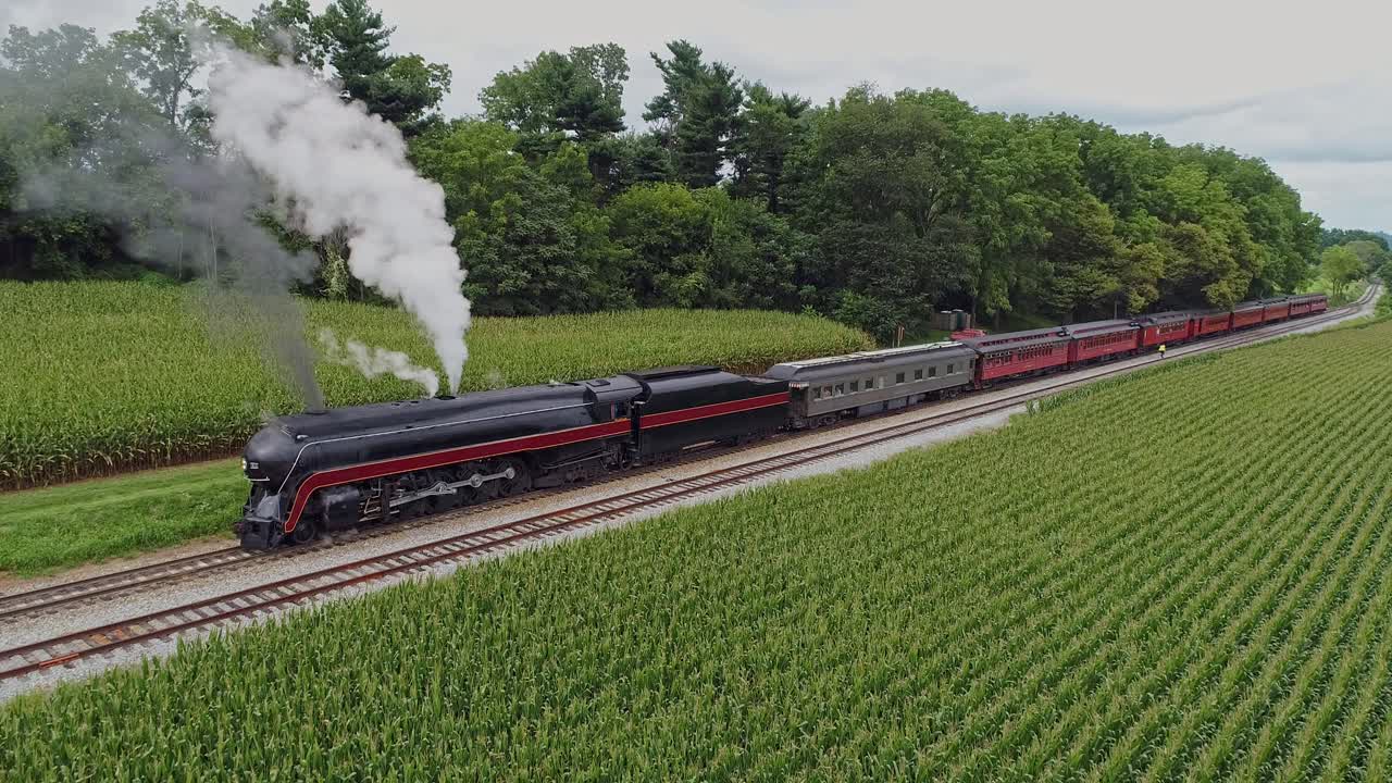 una vista aérea de frente a lado de un tren de pasajeros de vapor que sopla humo en una pequeña estación esperando a que los pasajeros aborden en un día de verano