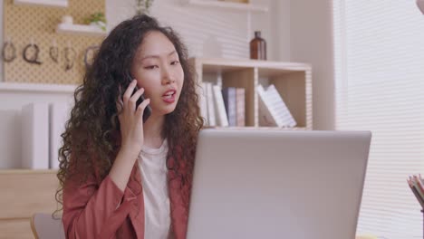 아시아 여성 디자이너는 스마트 폰을 사용하여 커뮤니케이션하고 프로젝트와 서류를 작업하며 노트북을 가진 책상에서 집에서 일합니다.