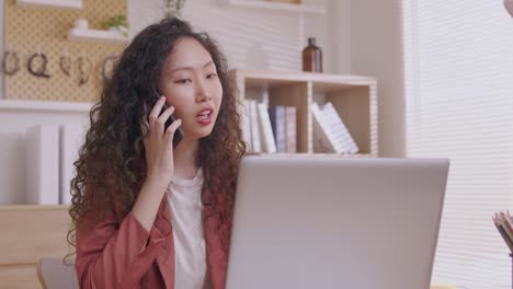 asian designer woman uses his smartphone to communicate. work on projects and papers. and work from home on a desk with a laptop.