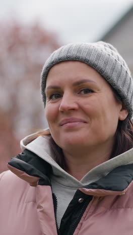 mujer confiada en chaqueta rosa y gorra gris con una sonrisa lúdica, de pie al aire libre contra el follaje borroso de otoño, casa y jardín de fondo