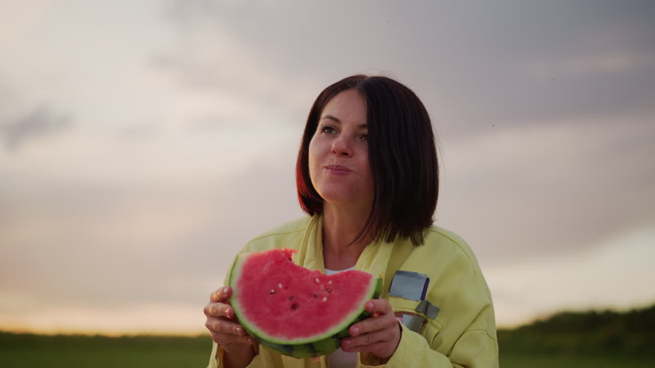 hora dorada saboreando sandía en un primer plano íntimo; una mujer sostiene una rodaja madura cerca de los labios, mordisqueando y sonriendo; pelo retroiluminado y cielo suave; campo tranquilo; restos de una cesta y una manta de picnic