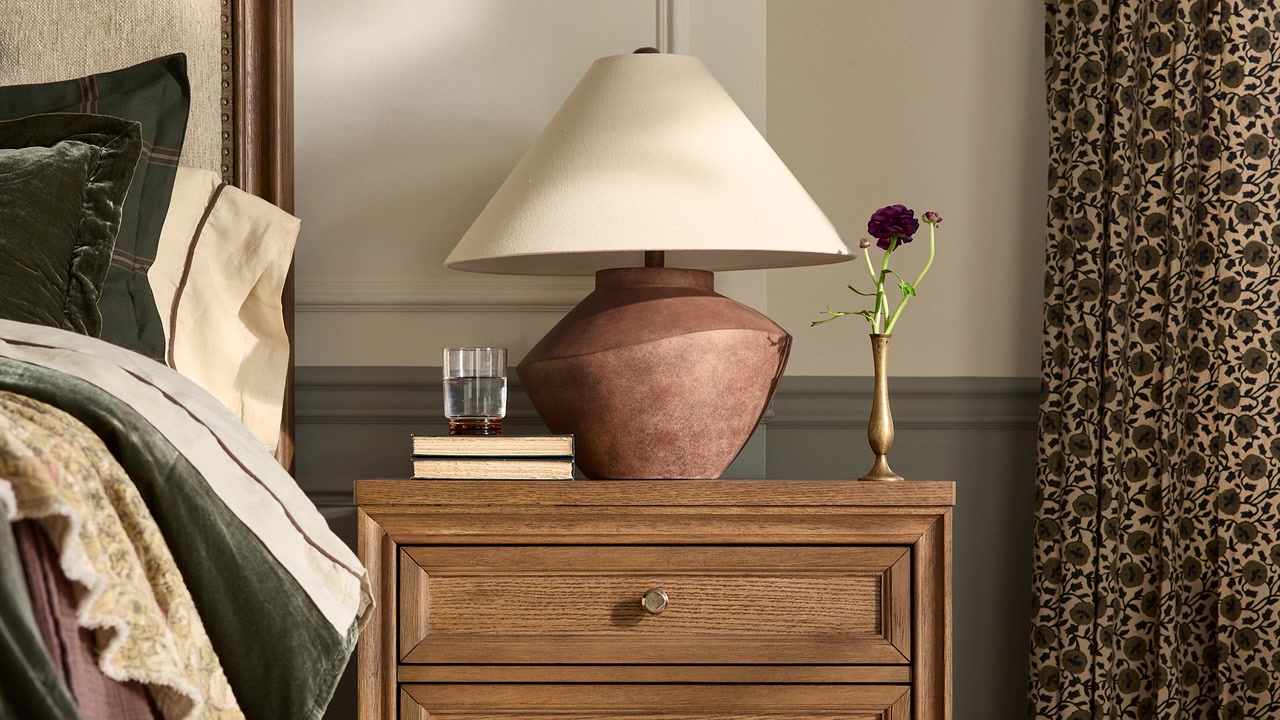 Slow zoom in on a farmhouse-style table lamp atop a rustic nightstand with flowers and a glass of water in a professionally staged bedroom.