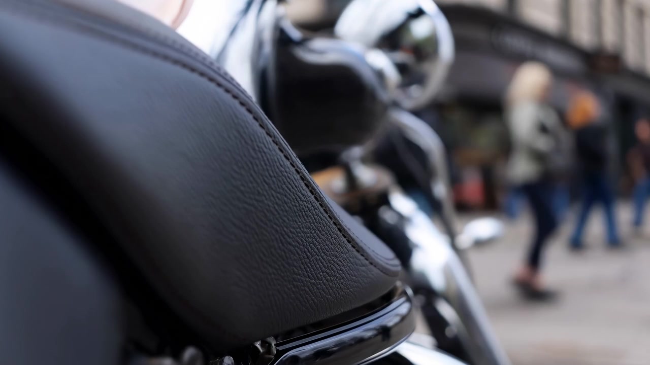 Primo piano dal basso di una moto parcheggiata, che si concentra sul sedile in pelle nera testurizzata in primo piano a sinistra e sul serbatoio lucido con forcella cromata e faro rotondo nelle vicinanze.