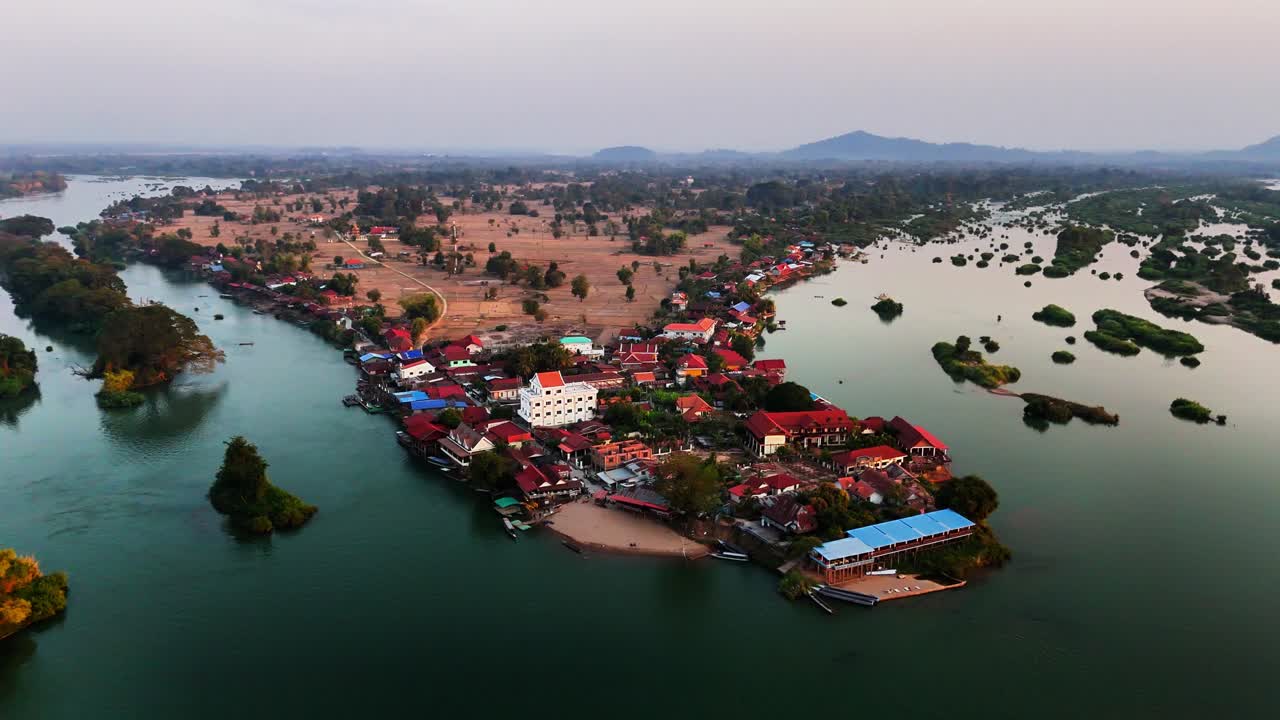 Drone view of Don Det in southern Laos featuring a riverside village, the branching Mekong River, boats, colorful rooftops, scattered islets, farmlands, and distant hills under soft evening light