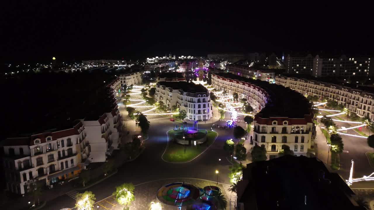 Nighttime aerial footage Phu Quoc brightly lit waterfront with European buildings along a dark waterway connected by illuminated pedestrian bridges and bustling walkways.